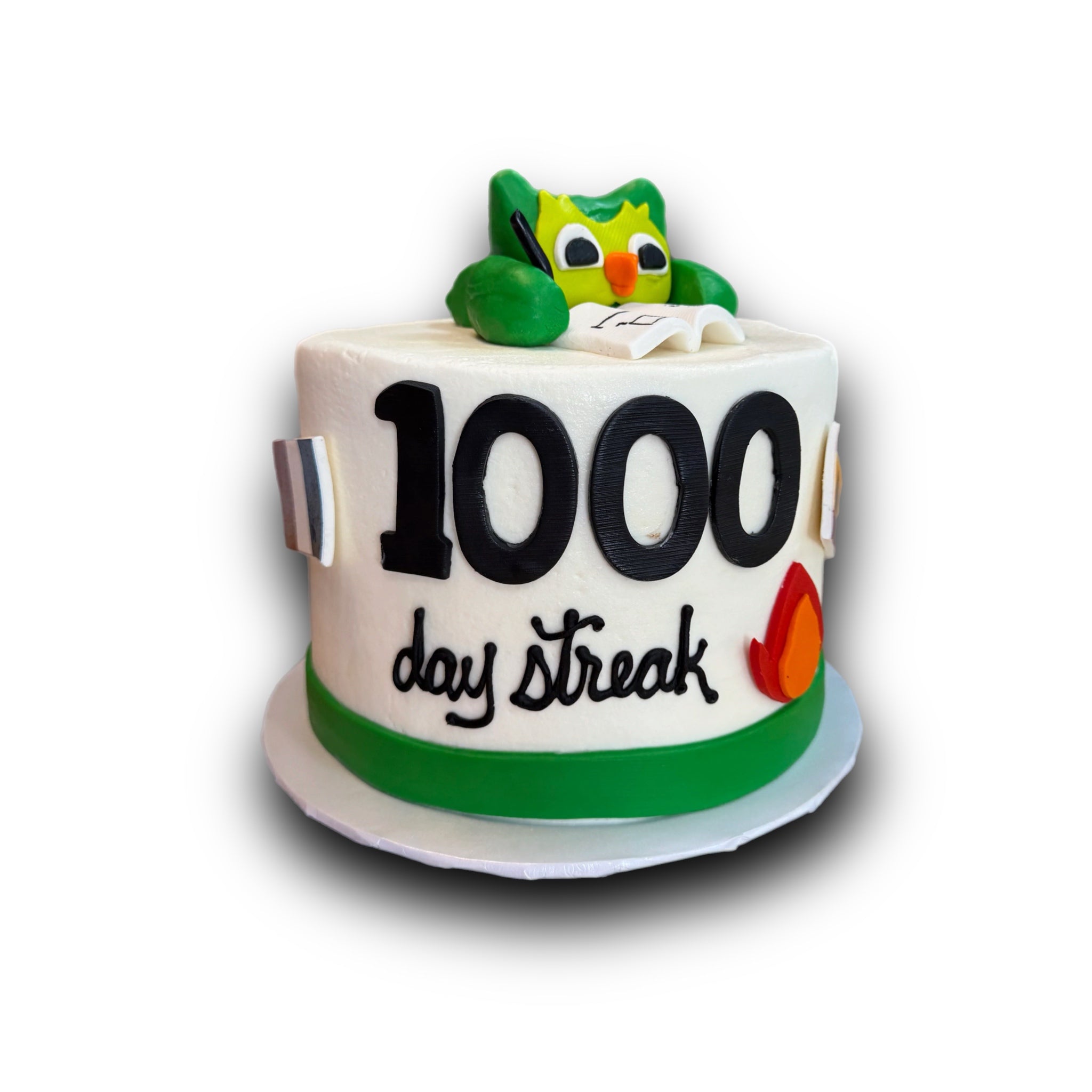 A white round birthday cake decorated with a green and yellow owl figurine, large black '1000' text, and 'day streak' in black script, with a green band around the lower middle, shown on a white background.