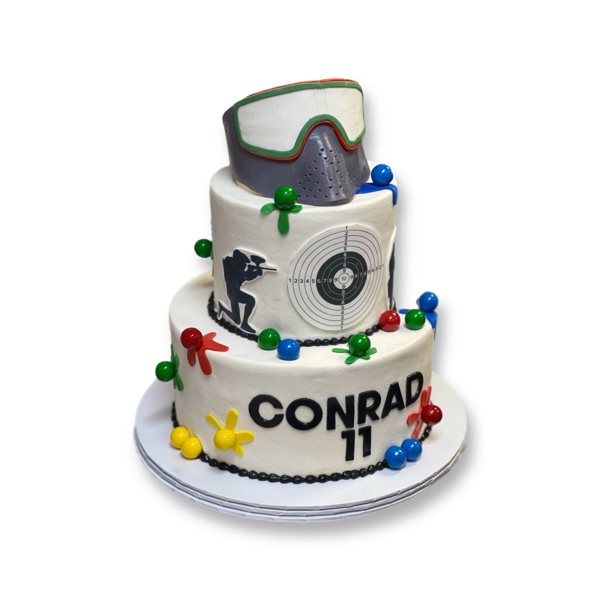 Two-tiered cake with a paint ball faskmask/helmet, target, and colorful decorations on a white background